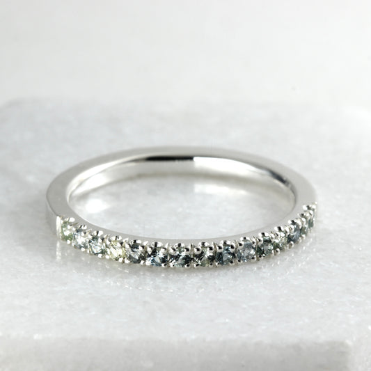 A sterling silver half eternity ring featuring a row of round-cut blue-gray and green Montana Sapphires, displayed on a light gray marble surface.
