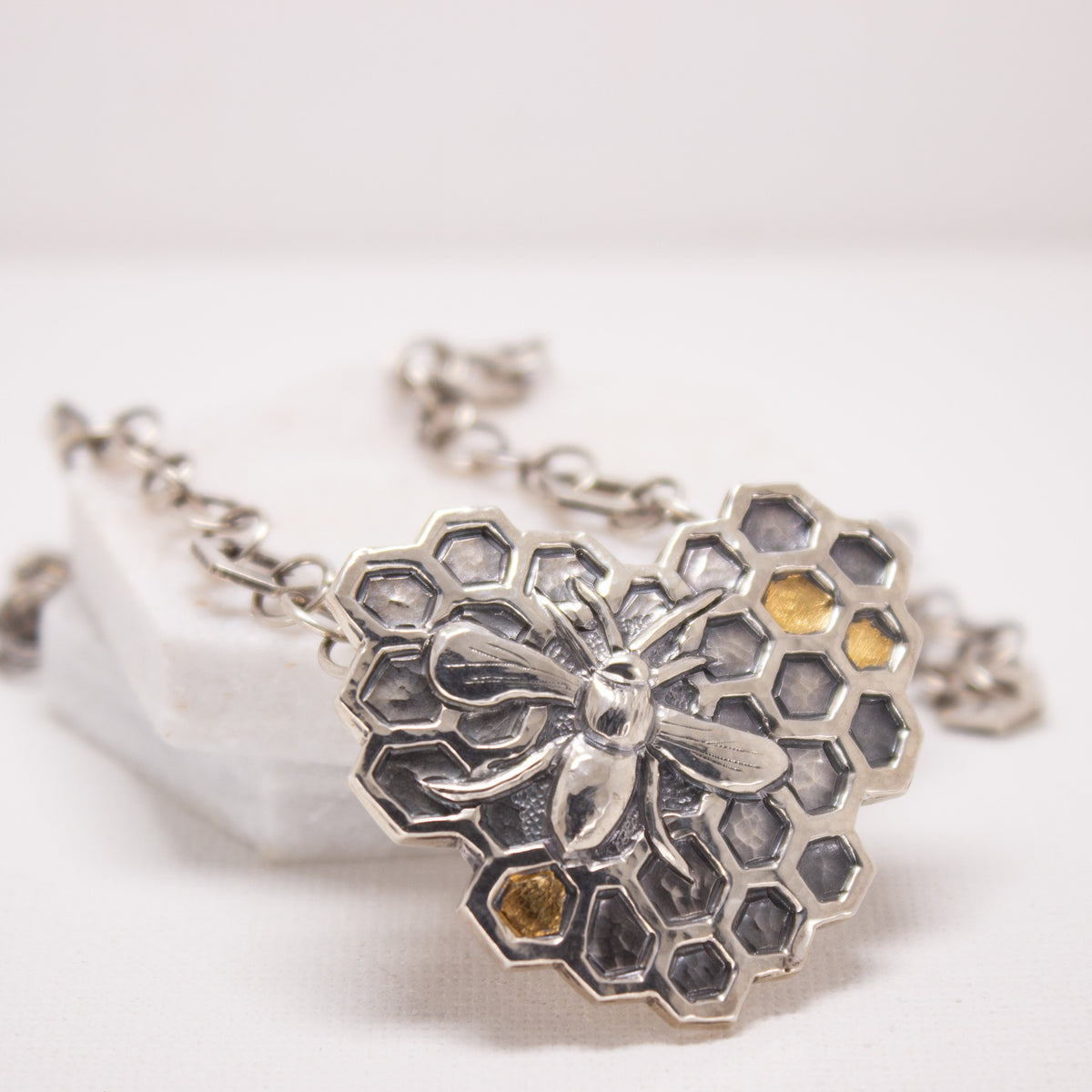 A sterling silver necklace shaped like a heart, filled with a honeycomb pattern and a bee. Some of the honeycomb has gold in it.