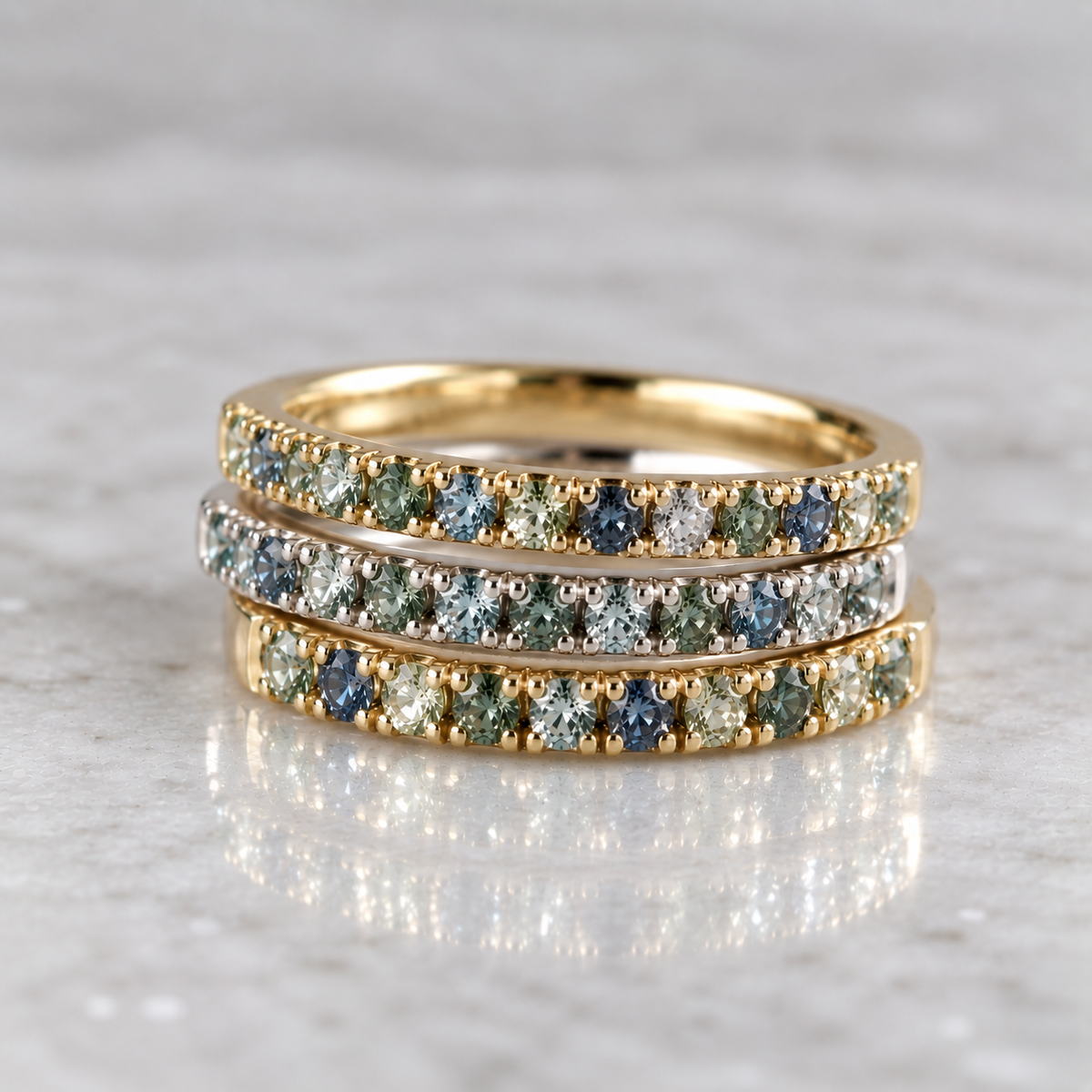 Set of three slender stacking rings, two in 14k gold and one in sterling silver, each set with blue and green Montana sapphires on a white marble surface.