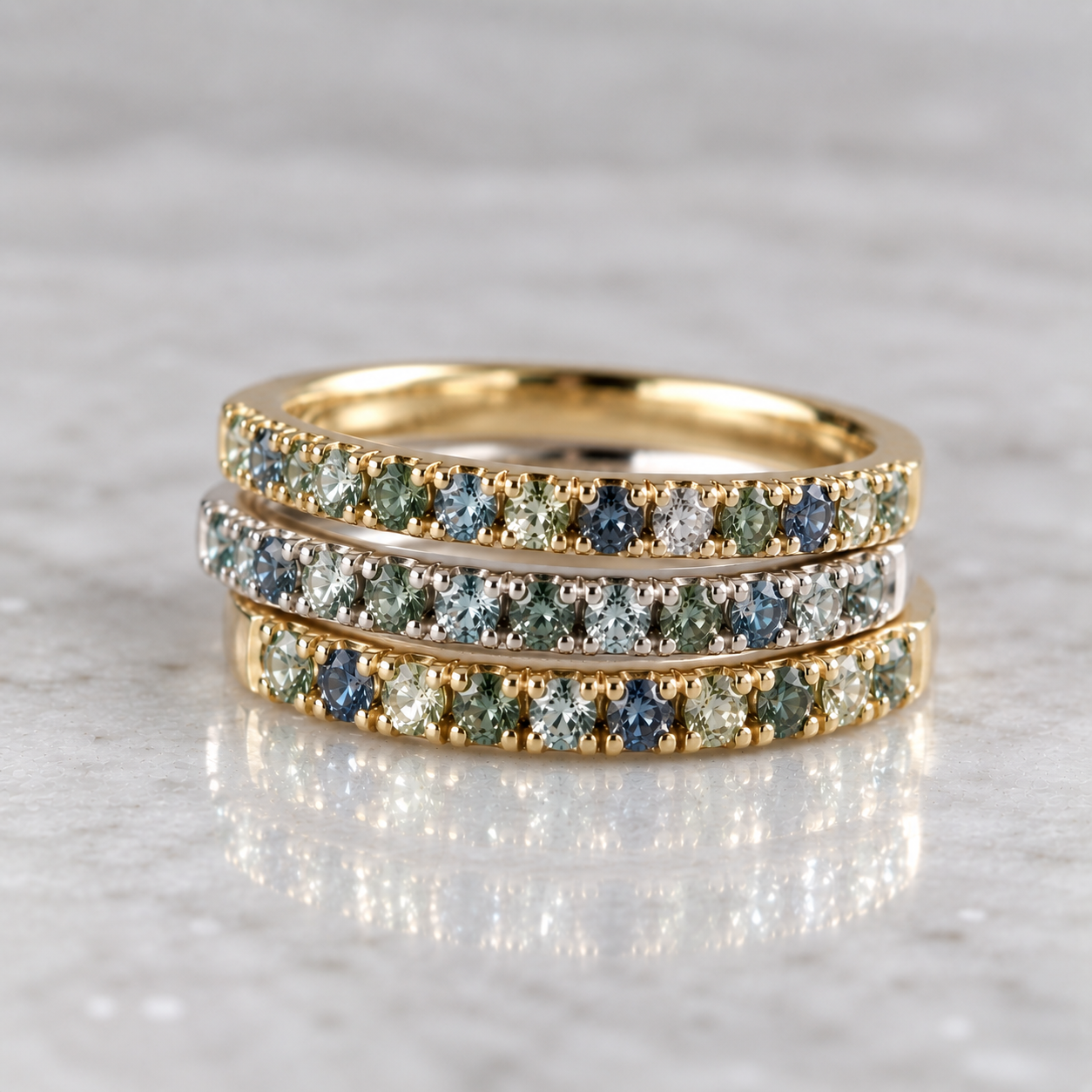 Set of three slender stacking rings, two in 14k gold and one in sterling silver, each set with blue and green Montana sapphires on a white marble surface.