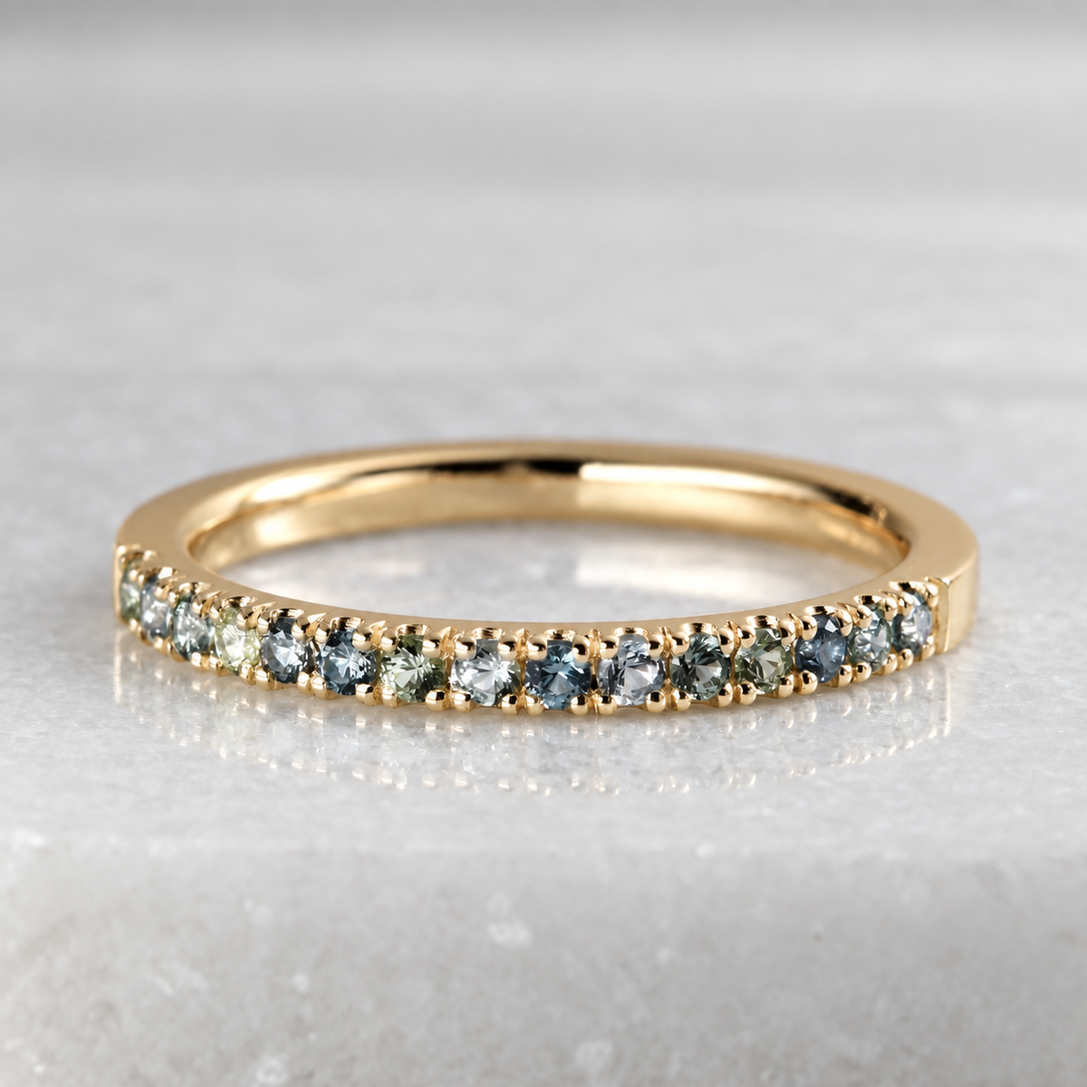 A yellow gold half eternity ring featuring a row of round-cut blue-gray and green Montana Sapphires, displayed on a light gray marble surface.
