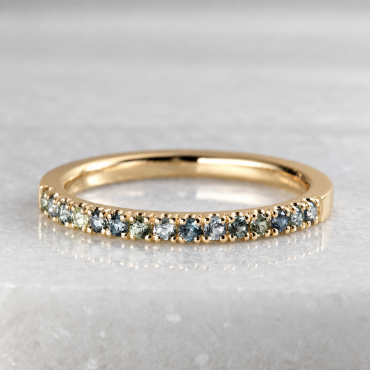 A yellow gold half eternity ring featuring a row of round-cut blue-gray and green Montana Sapphires, displayed on a light gray marble surface.