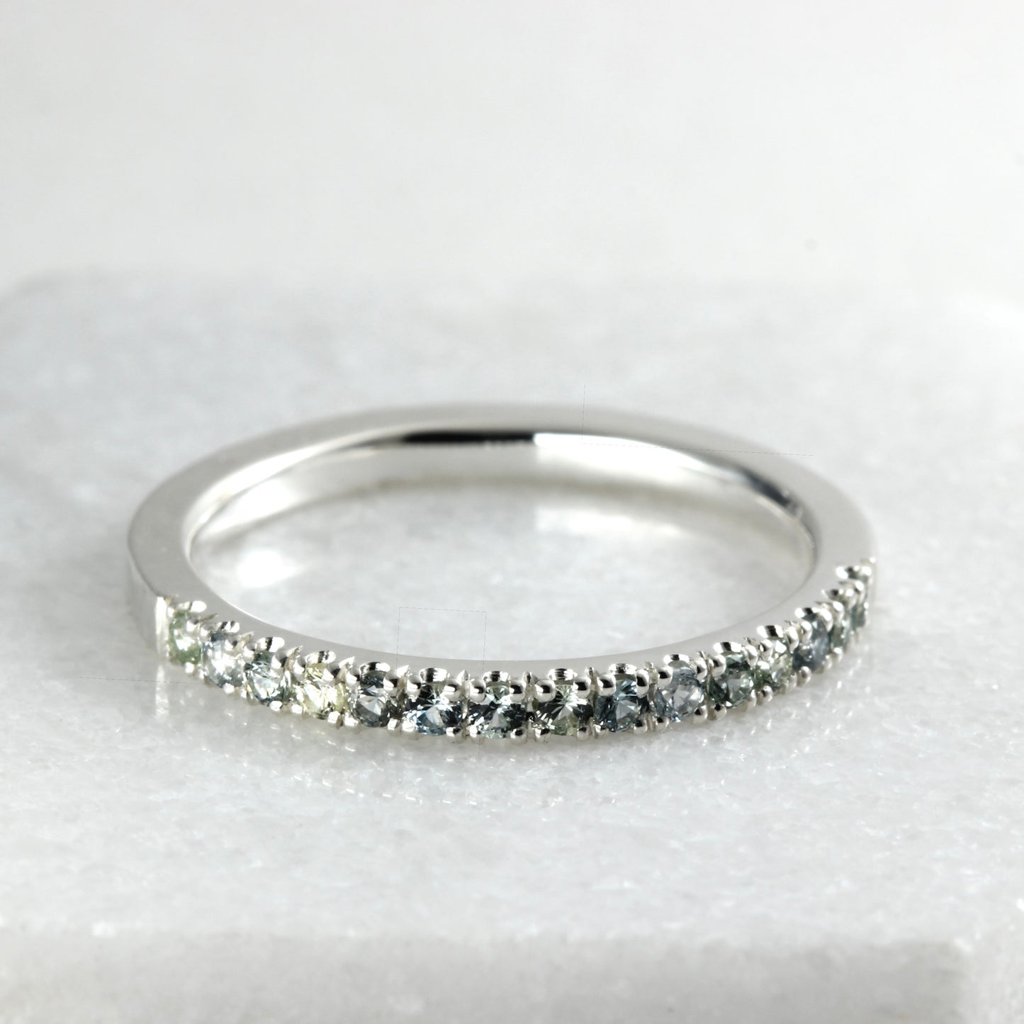 A sterling silver half eternity ring featuring a row of round-cut blue-gray and green Montana Sapphires, displayed on a light gray marble surface.