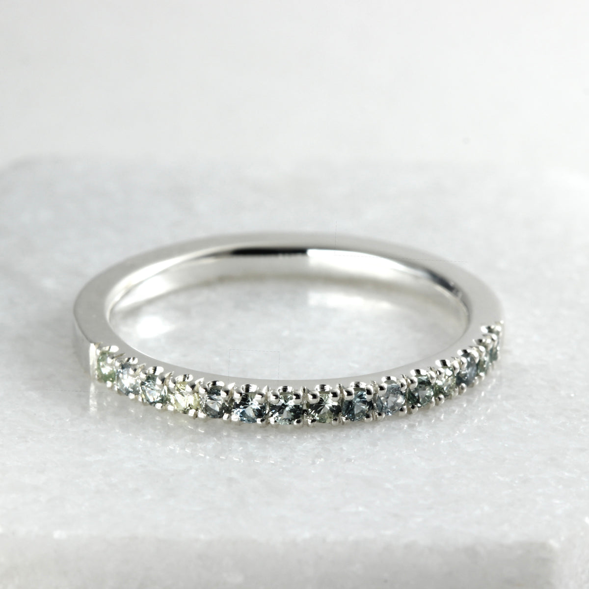 A sterling silver half eternity ring featuring a row of round-cut blue-gray and green Montana Sapphires, displayed on a light gray marble surface.