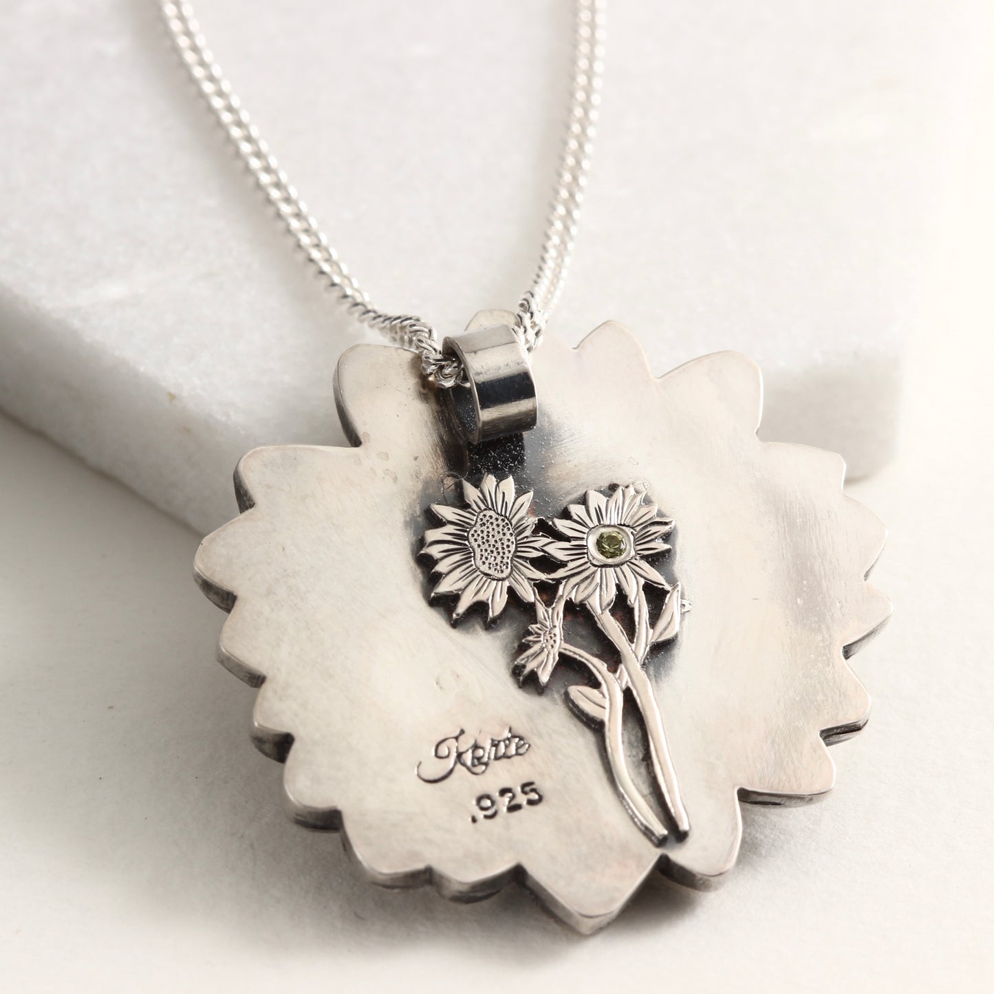 Back side of Wild Radiance Daisy Pendant with gold center in sterling silver on white marble background, showing engraved daisy with Montana sapphire